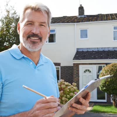 Man with grey hair and beard smiles at the camera while holding a clipboard in front of a home, presumably exhibiting the best course of action after a home inspection.
