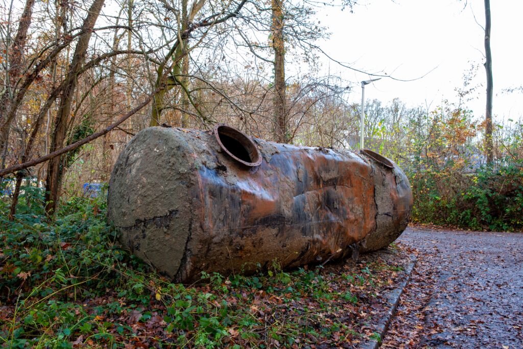 Rusted, old oil tank that has been removed from someone’s soil and placed next to trees.