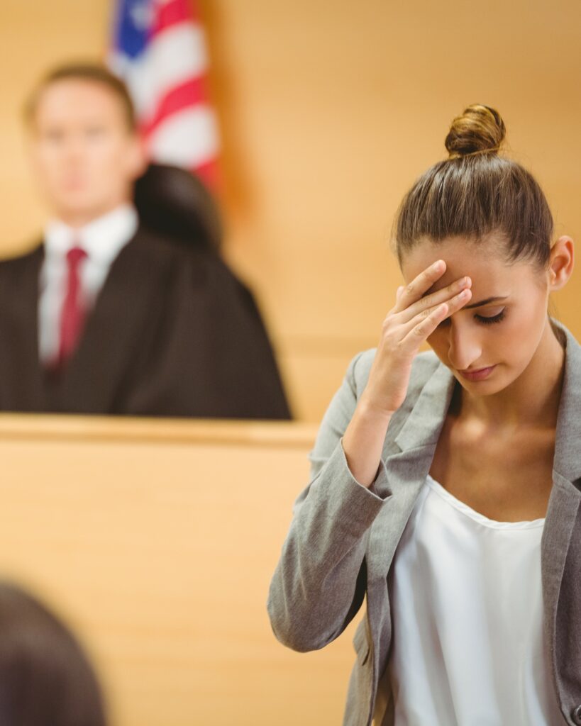 Woman stands before a judge in a courtroom holding her face with one hand, signifying stress as one might in a home inspector court summons or subpoena.