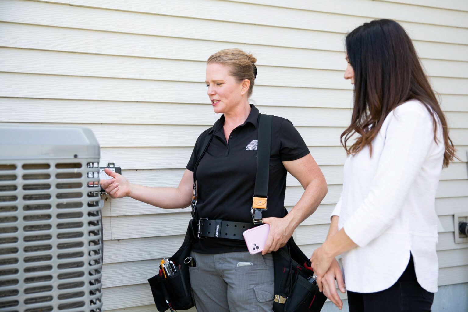 Woman wearing a toolbelt inspects an air conditioning unit and explains her home inspection findings to a realtor or client. Photo courtesy of Structure Tech.