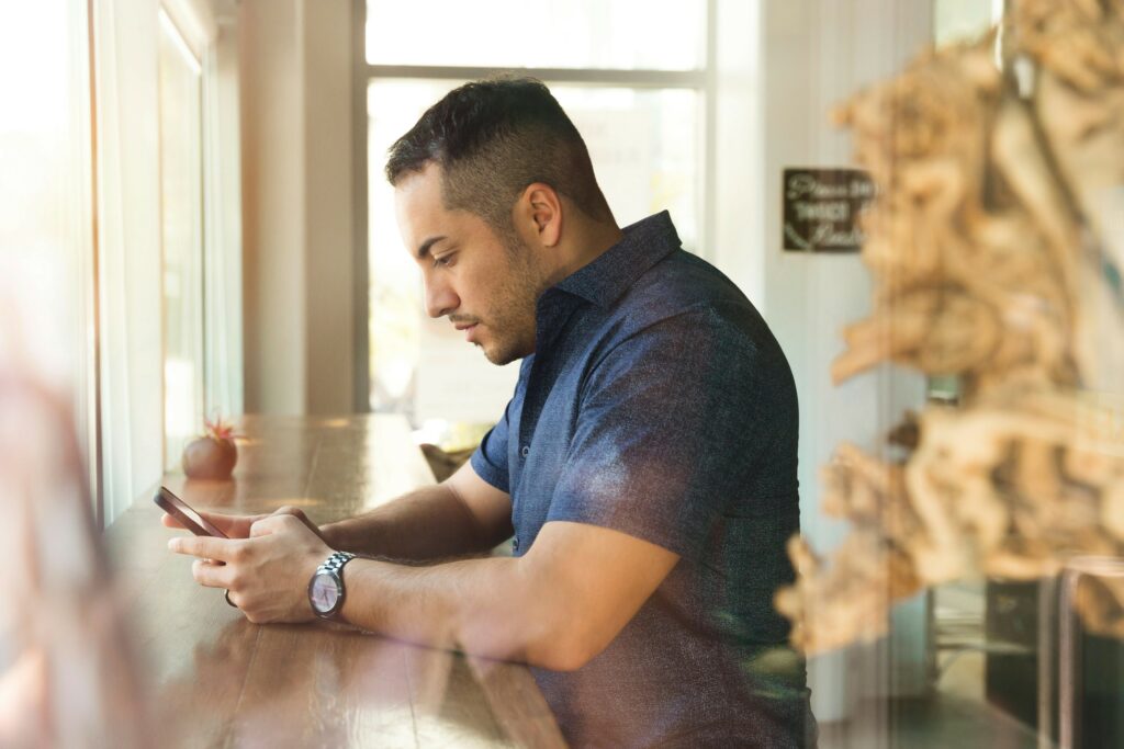 Man in a blue collared shirt sits at a counter looking down at his phone, presumably in a coffee shop, practicing timely claims reporting.