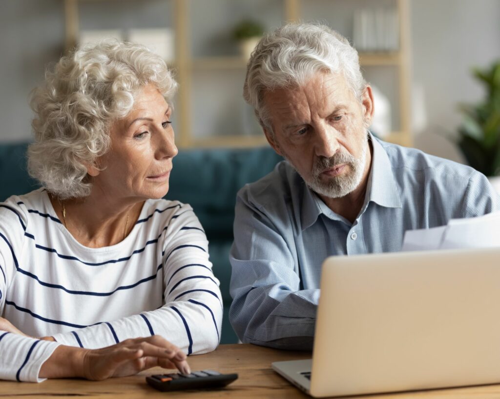Elderly man and woman discuss documents, like home inspection contracts, together while sitting at a table in front of a laptop.