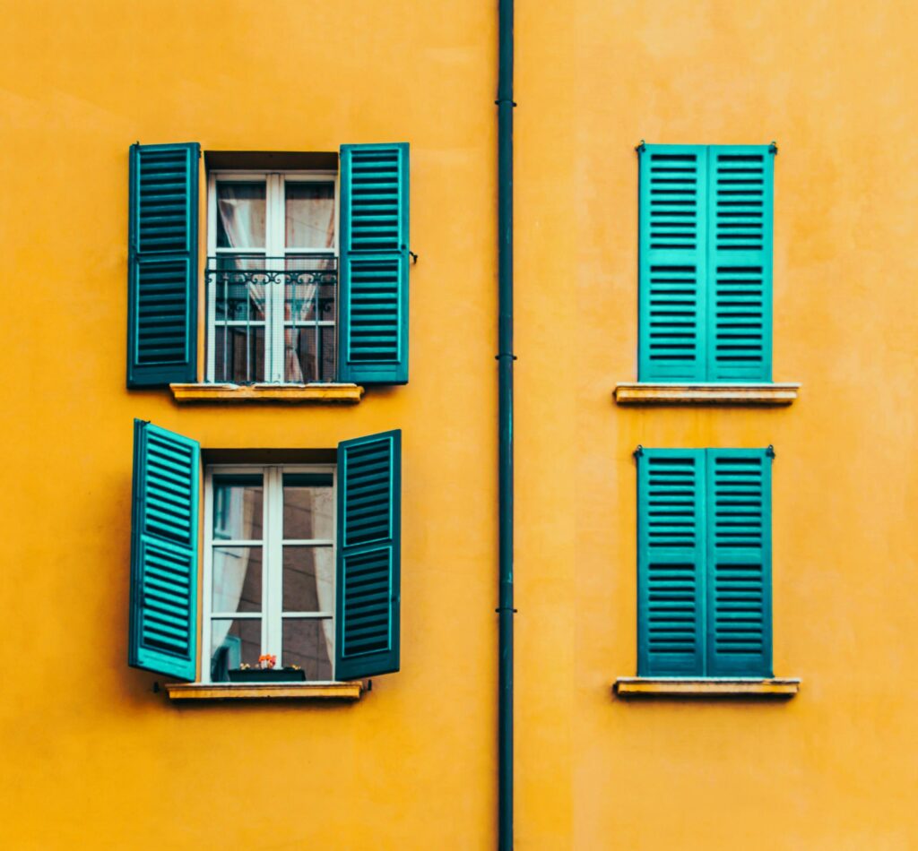 Yellow house with teal wooden window panes opening to the outside, four windows lined up in a square.