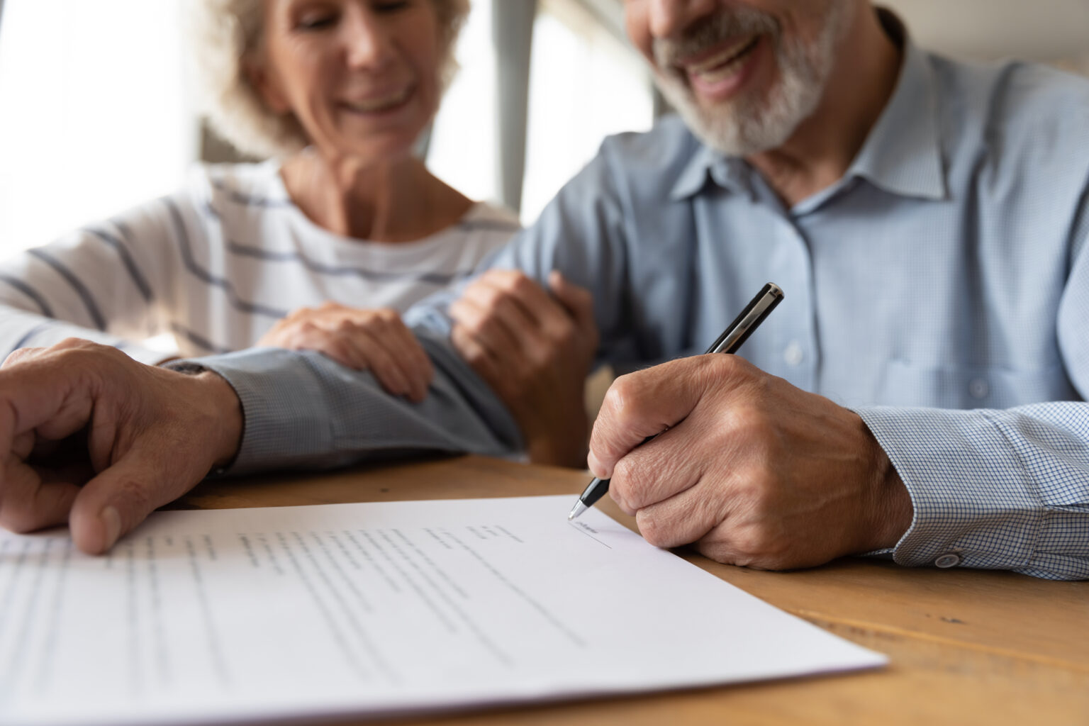Zoomed-in image of elderly man and woman sitting together and signing a home inspection contract on paper, smiles on their faces.