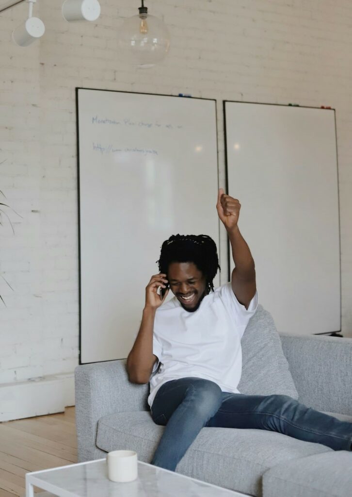 Man in t-shirt and jeans sits on a couch with a phone pressed to his face, making an excited expression as one who is excited about starting a home inspection business.