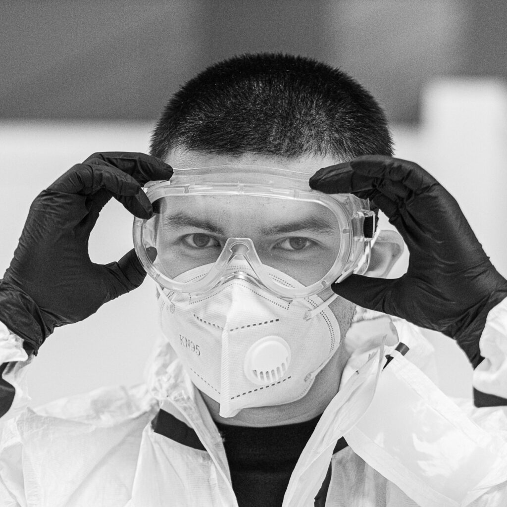 Greyscale of home inspector putting on eye protection safety and ppe eye protection goggles, while wearing gloves and an air filter mask, presumably to enter a dirty crawlspace.