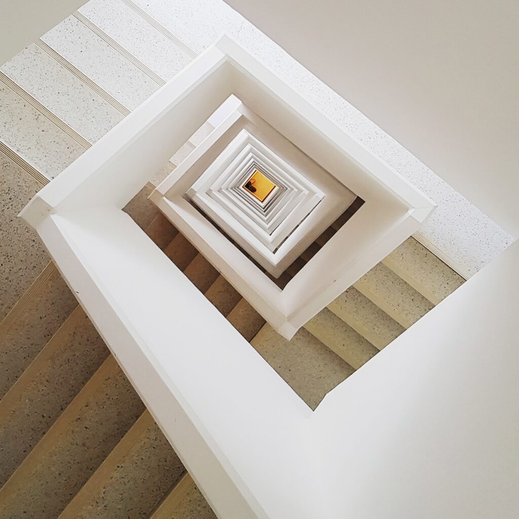 Camera panned down an indoor staircase. The staircase has white walls, tan steps, and descends in a square-shaped spiral for several flights. The image suggests an indoor staircase and common area for apartments and condos.