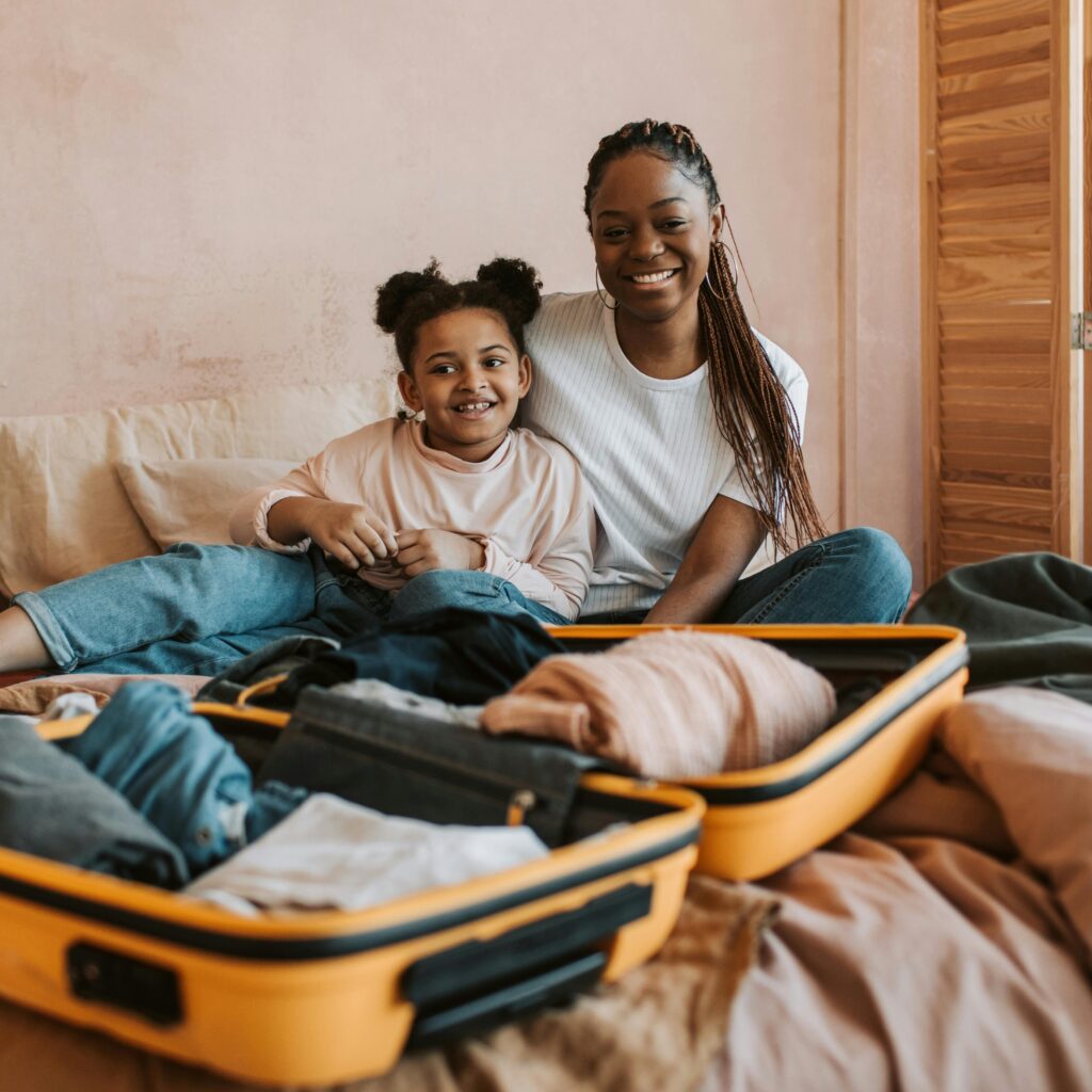 Smiling mother and daughter sitting on a vacation home bed next to an open suitcase.