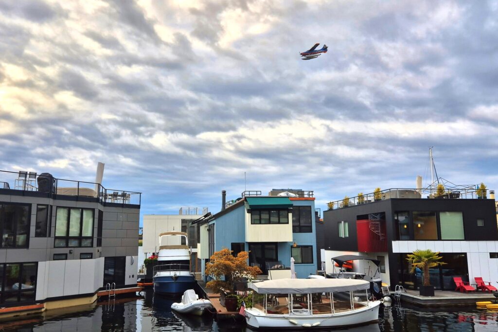 A floating home community in Seattle with multiple, colorful floating houses, anchored boats, and a small plane flying overhead.