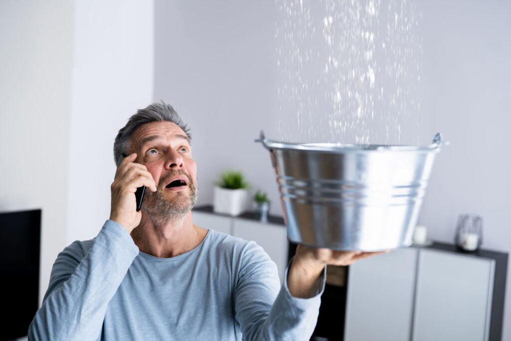 Man holds out a metal bucket with one hand to catch water dripping from the ceiling, and holds phone to his ear with the other hand, a shocked expression on his face.